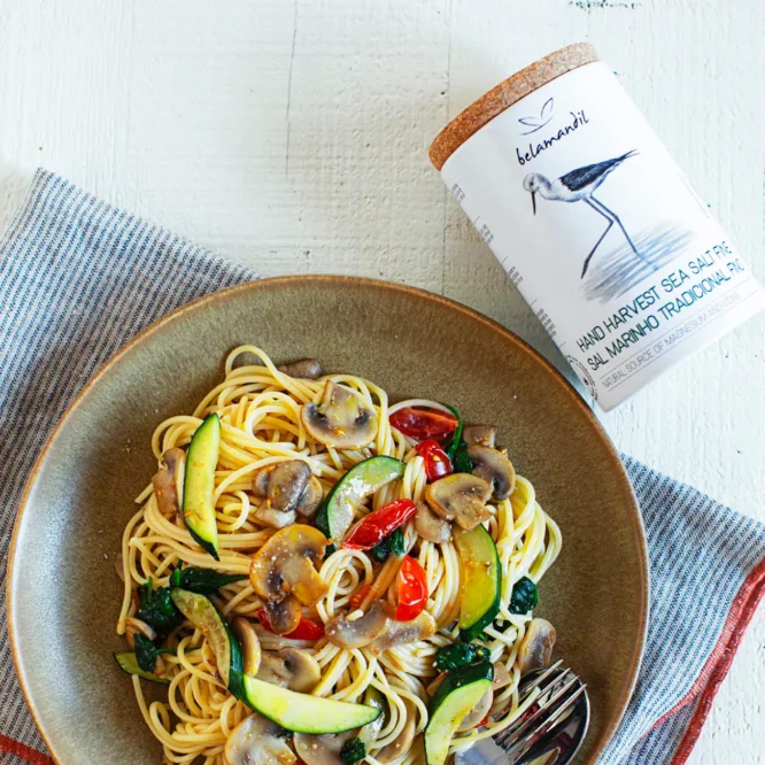 Plate of pasta with vegetables and a container of Belamendi sea salt on a white surface.