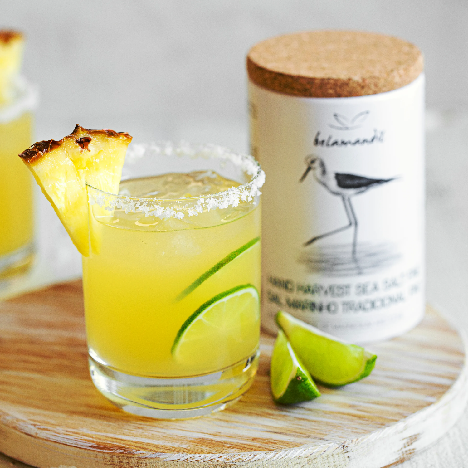 Glass of yellow cocktail with pineapple wedge and lime on a wooden board with a bottle in the background.