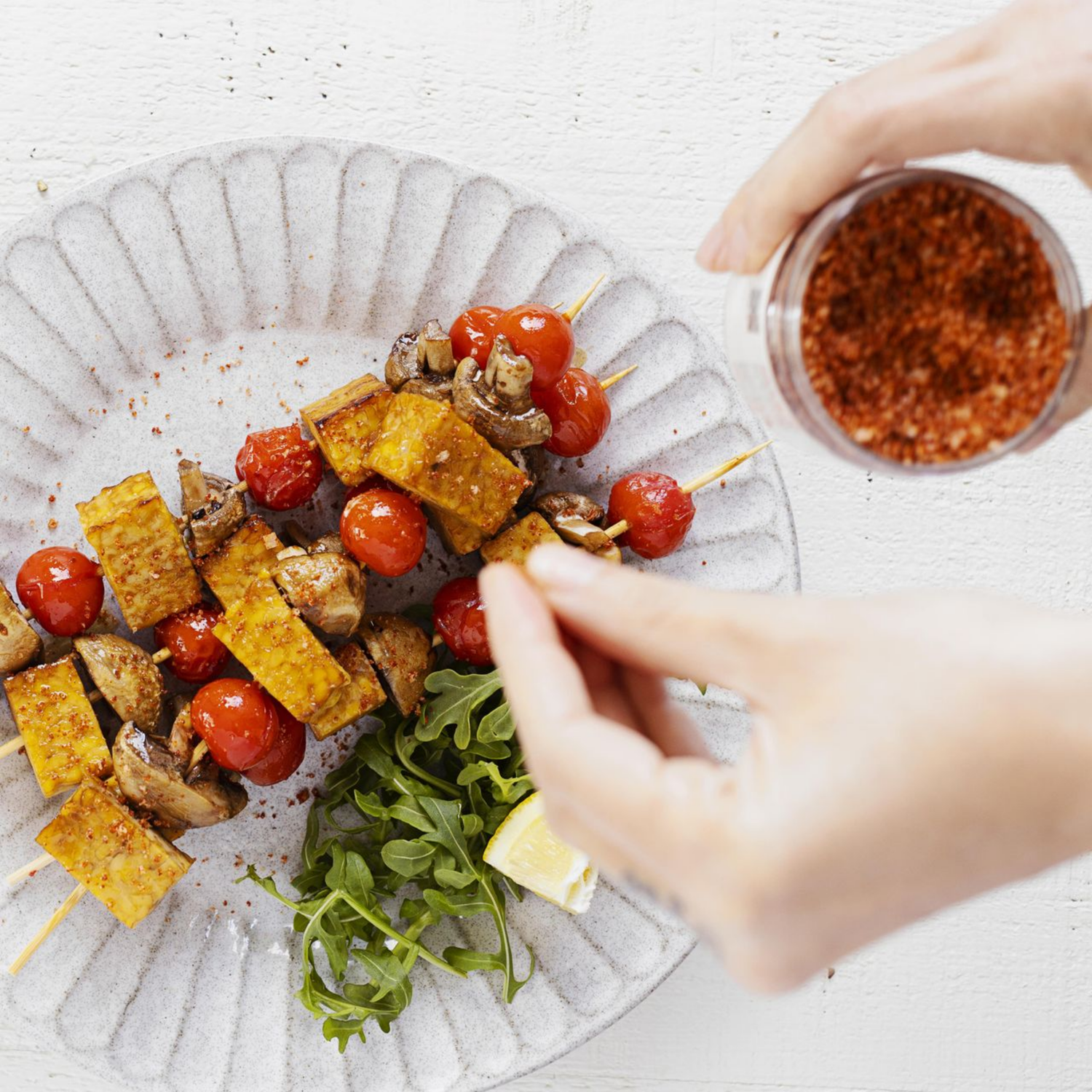 Person seasoning skewers of tofu and cherry tomatoes with a spice container on a white surface.