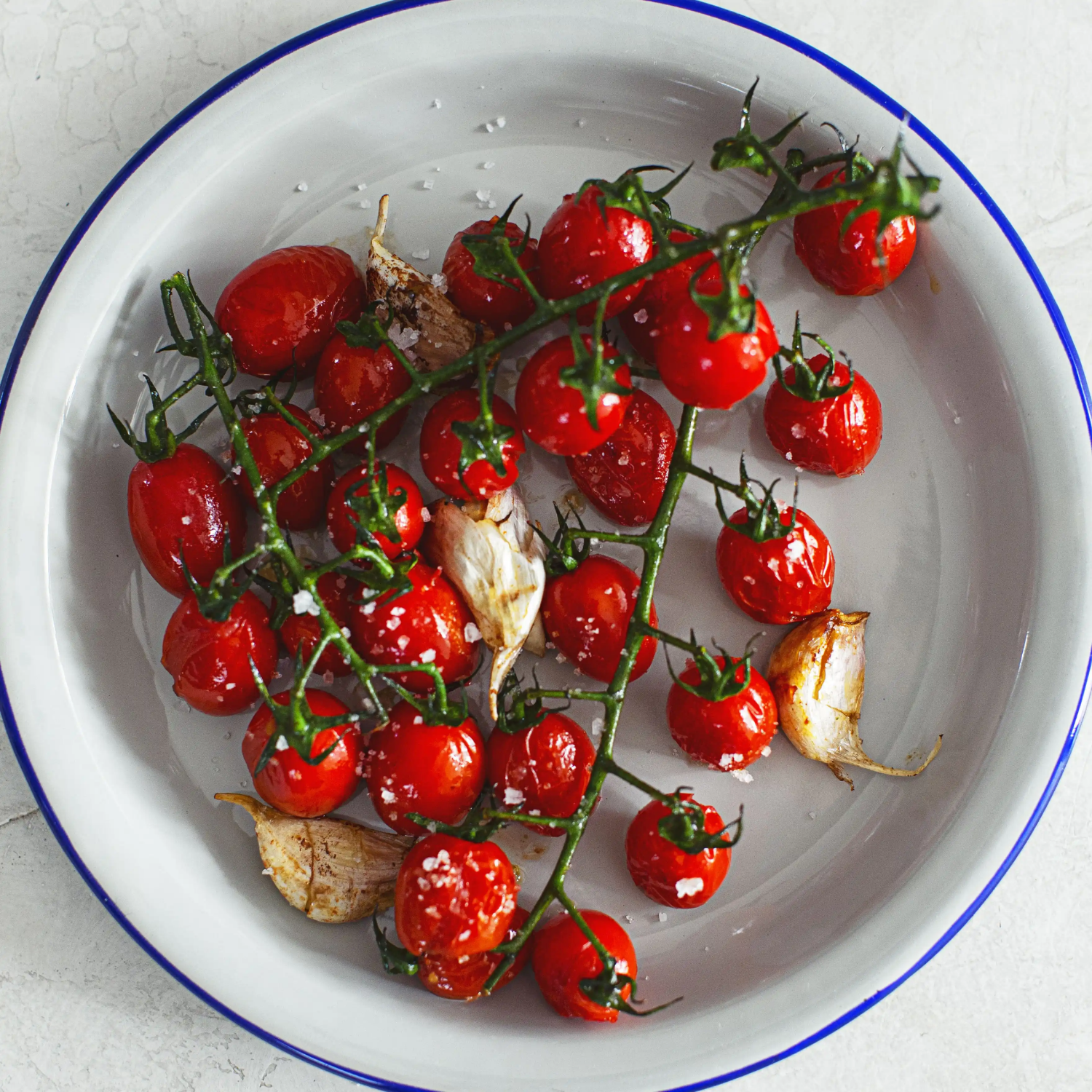Roasted cherry tomatoes on a vine with garlic cloves in a white bowl with blue rim.