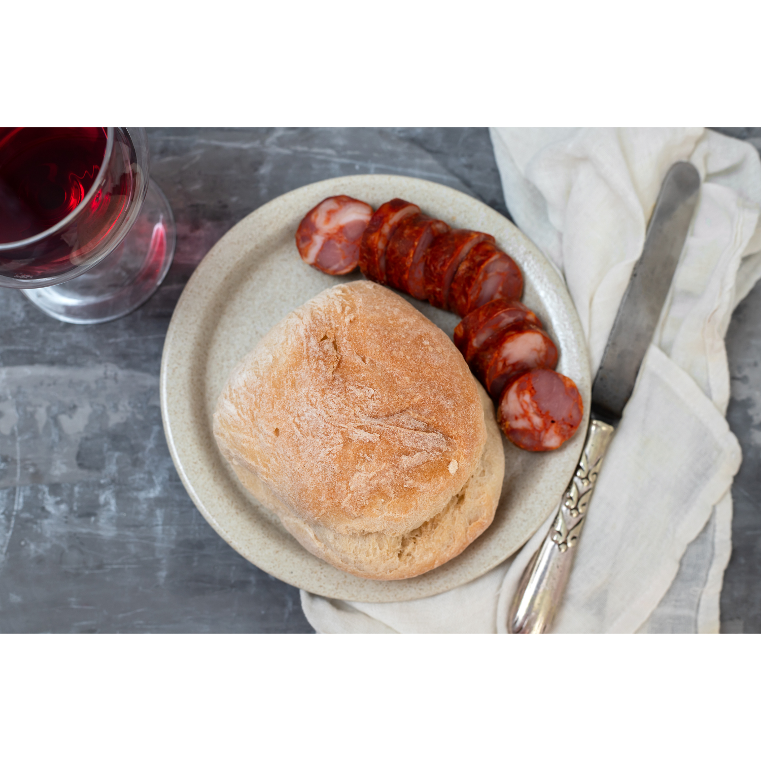 Chouriço caseiro slices on a plate with a rustic bread roll and a glass of red wine beside it.