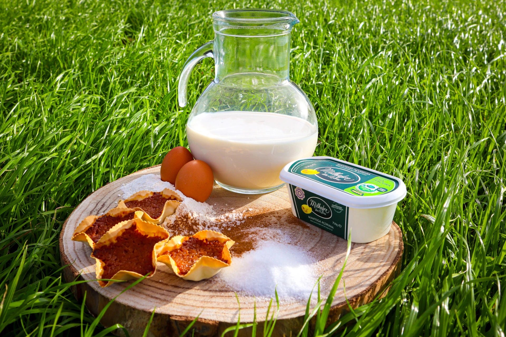 Queijadas da Graciosa on a wooden board with milk, eggs, sugar, and butter, set against a grassy background.